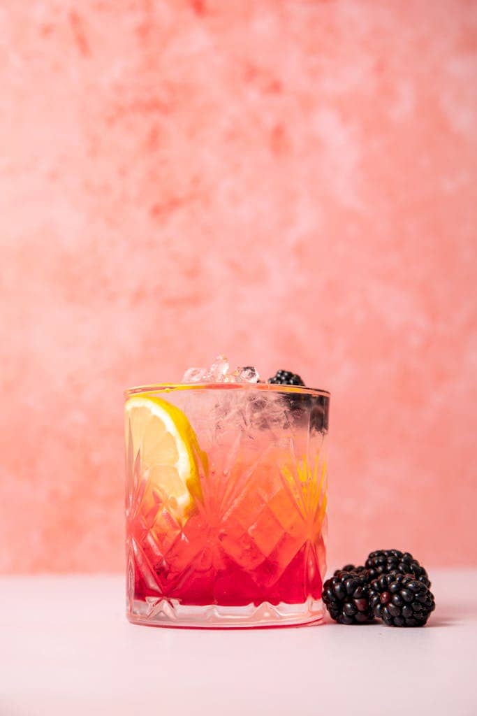 A vibrant blackberry and lemon cocktail in a crystal glass with a pink backdrop.
